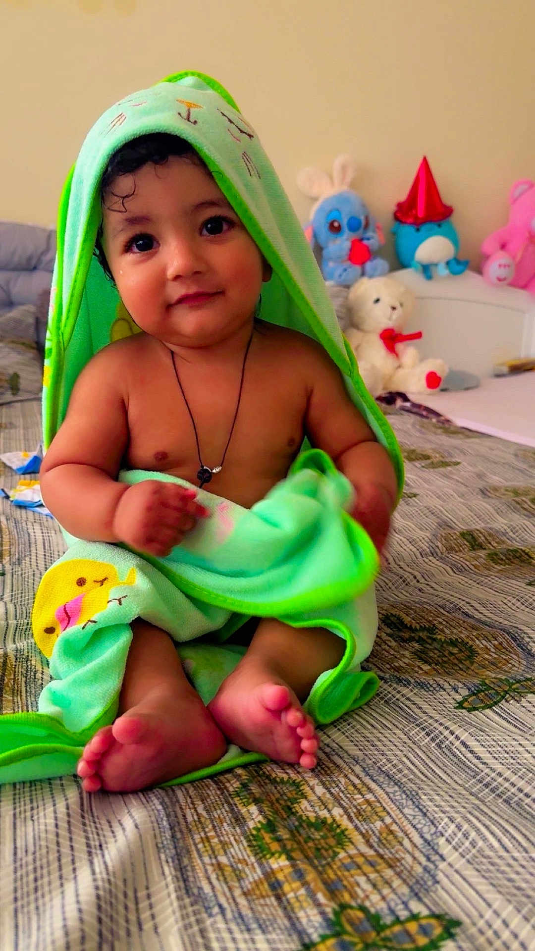 Kevin is registered to the contest to win money with this photo: baby, child, towel, bed, stuffed_animals, toy, plush, cute, indoors, smile, face, feet, necklace, hood, patterned_bedspread, sitting, infant, soft_toy, colorful, cozy