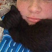 Rocket joined the competition — help win amazing prizes! person, kitten, black_cat, face, blue_shirt, text_on_shirt, crocheted_blanket, pillow, floral_pattern, close_up, indoor, lying_down, pet, cute, cozy, resting, animal, young, casual, portrait