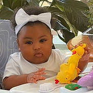 Dalanii is registered to the contest to win money with this photo: baby, child, high_chair, white_bow, toy, yellow_toy, indoor, plants, greenery, curious, infant, headband, furniture, table, hand, foot, cute, person, seated, home