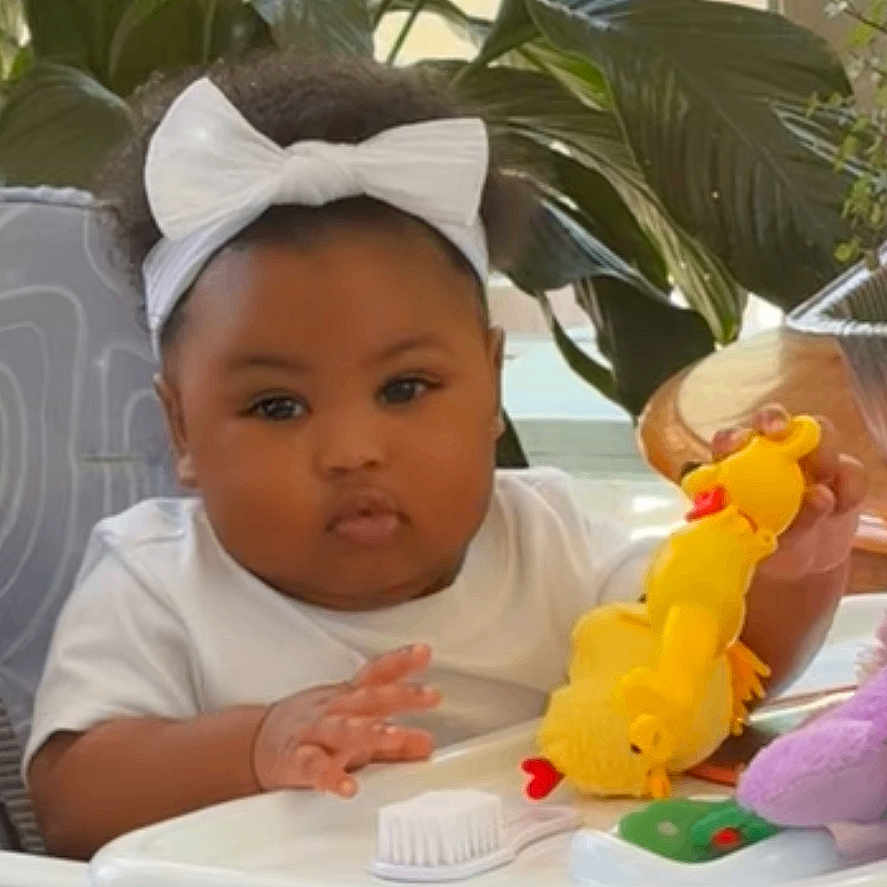 Dalanii is registered to the contest to win money with this photo: baby, child, curious, cute, foot, furniture, greenery, hand, headband, high_chair, home, indoor, infant, person, plants, seated, table, toy, white_bow, yellow_toy