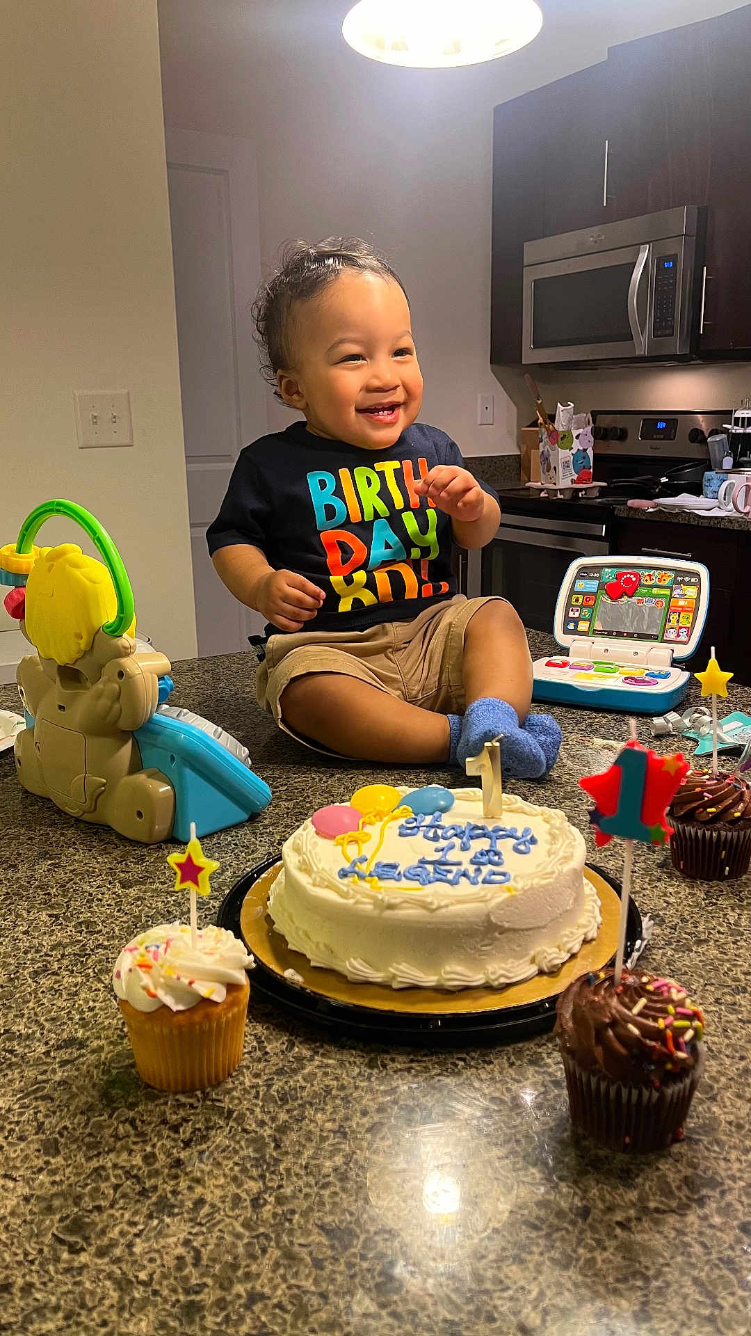 Legend is registered to the contest to win money with this photo: toddler, birthday, cake, cupcake, toy, kitchen, counter, smiling, child, celebration, candle, clothing, furniture, indoor, food, person, happy, toy_laptop, cup, lighting