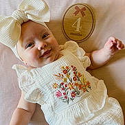 Blakley is registered to the contest to win money with this photo: baby, infant, smiling, portrait, lying_down, white_bow, headband, dress, embroidered, floral, wooden_plaque, milestone, four_months, indoor, soft_background, textured_blanket, close_up, happy, child, photography
