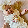 baby, infant, smiling, portrait, lying_down, white_bow, headband, dress, embroidered, floral, wooden_plaque, milestone, four_months, indoor, soft_background, textured_blanket, close_up, happy, child, photography