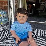 Hayes joined the competition — help win amazing prizes! toddler, child, sitting, blue_shirt, shorts, indoor_outdoor, patterned_rug, face, curious_eyes, hands, barefoot, water_bottles, brick_wall, doorway, man, sign, text, furniture, floor, casual