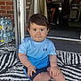 toddler, child, sitting, blue_shirt, shorts, indoor_outdoor, patterned_rug, face, curious_eyes, hands, barefoot, water_bottles, brick_wall, doorway, man, sign, text, furniture, floor, casual