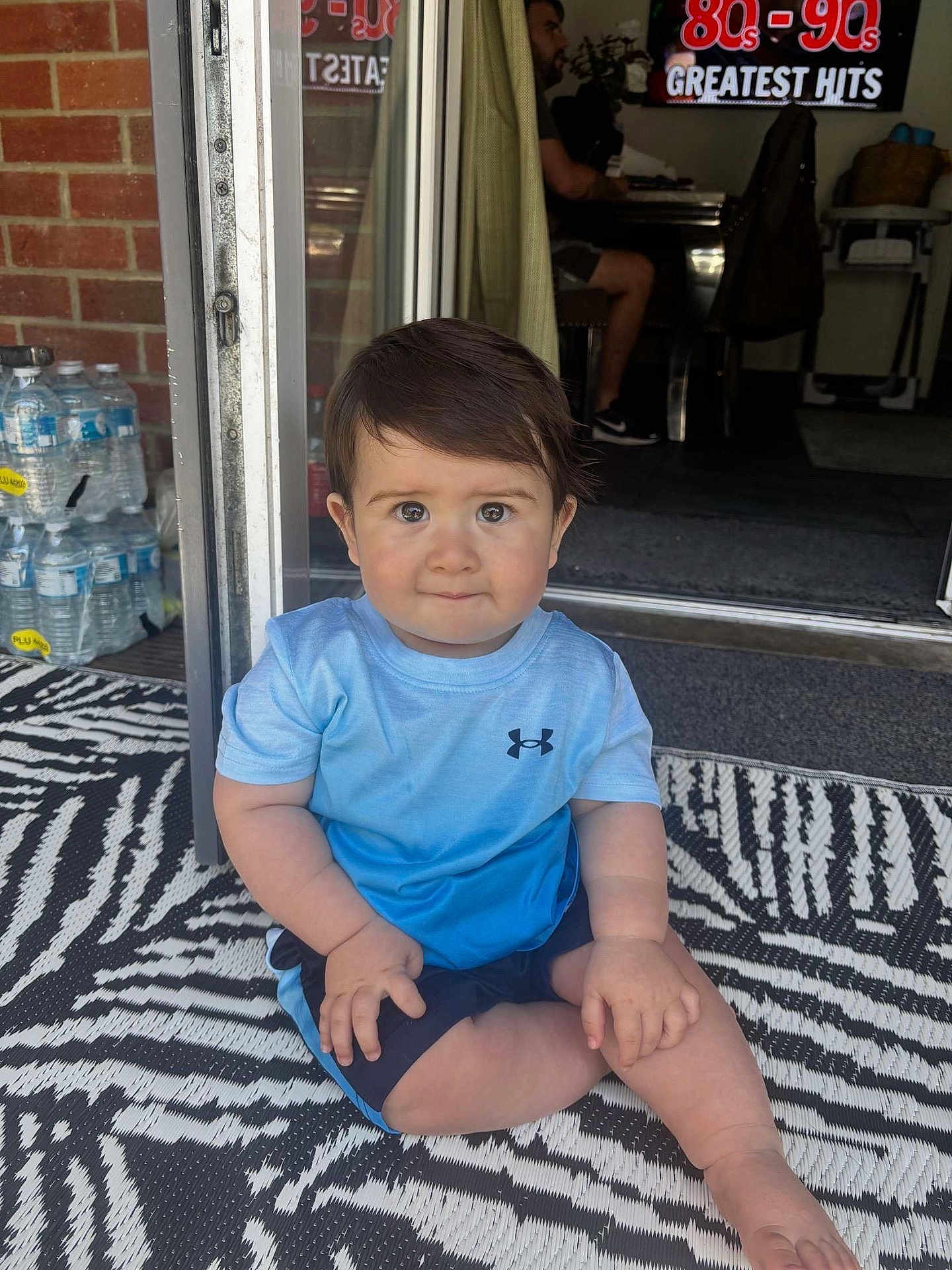 Hayes joined the competition — help win amazing prizes! toddler, child, sitting, blue_shirt, shorts, indoor_outdoor, patterned_rug, face, curious_eyes, hands, barefoot, water_bottles, brick_wall, doorway, man, sign, text, furniture, floor, casual