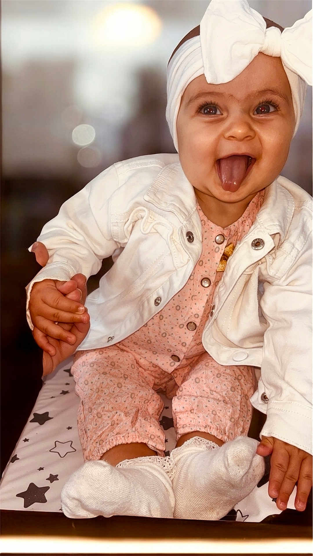 Alba participe au concours pour gagner de l'argent avec cette photo : baby, child, tongue_out, white_jacket, pink_outfit, headband, bow, sitting, holding_hand, smiling, happy, indoor, changing_pad, stars_pattern, sock, cute, portrait, infant, playful, hand