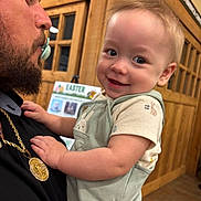 Waylon is registered to the contest to win money with this photo: baby, child, man, beard, pacifier, smile, holding, indoor, wooden_door, sign, easter, clothing, overalls, portrait, happy, person, face, hand, background, floor