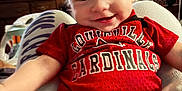 Waylon is registered to the contest to win money with this photo: baby, child, smiling, red_shirt, louiville_cardinals, person, indoor, furniture, television, home, legs, pants, sitting, face, happy, casual, cozy, background, people, clothing
