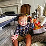 baby, child, infant, smiling, sitting, playpen, plush_toy, giraffe, elephant, stuffed_animal, toys, nursery, fireplace, chair, blanket, pajamas, plaid_shirt, indoor, portrait, happy