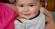 Sebastian joined the competition — help win amazing prizes! adult_hand, baby, blue_eyes, caregiver, child, close_up, clothing, face, family, happy, home, indoors, infant, pink_clothing, portrait, seated, short_hair, smiling, striped_shirt, wood_paneling