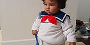 Draven is registered to the contest to win money with this photo: toddler, child, costume, sailor_outfit, hat, red_bow, white_clothing, curly_hair, indoor, floor, tile_floor, toy, blue_spatula, standing, pouty_expression, person, young_child, clothing, home, curious