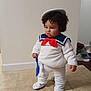 toddler, child, costume, sailor_outfit, hat, red_bow, white_clothing, curly_hair, indoor, floor, tile_floor, toy, blue_spatula, standing, pouty_expression, person, young_child, clothing, home, curious