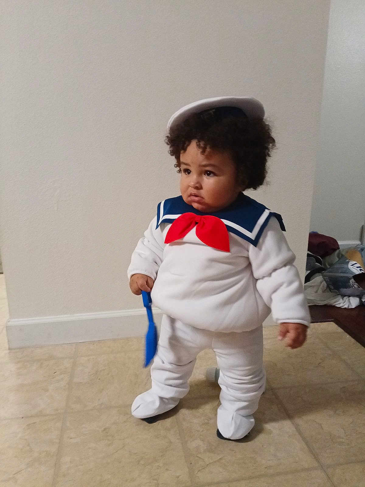 Draven is registered to the contest to win money with this photo: toddler, child, costume, sailor_outfit, hat, red_bow, white_clothing, curly_hair, indoor, floor, tile_floor, toy, blue_spatula, standing, pouty_expression, person, young_child, clothing, home, curious