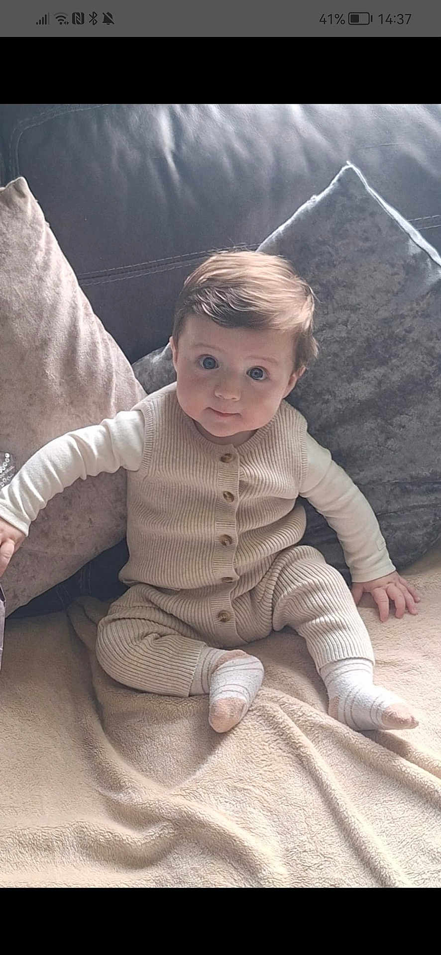 Rocco is registered to the contest to win money with this photo: baby, child, infant, knitwear, beige, socks, blanket, couch, pillows, face, blue_eyes, cute, sitting, indoors, soft_texture, home, comfort, young, person, portrait