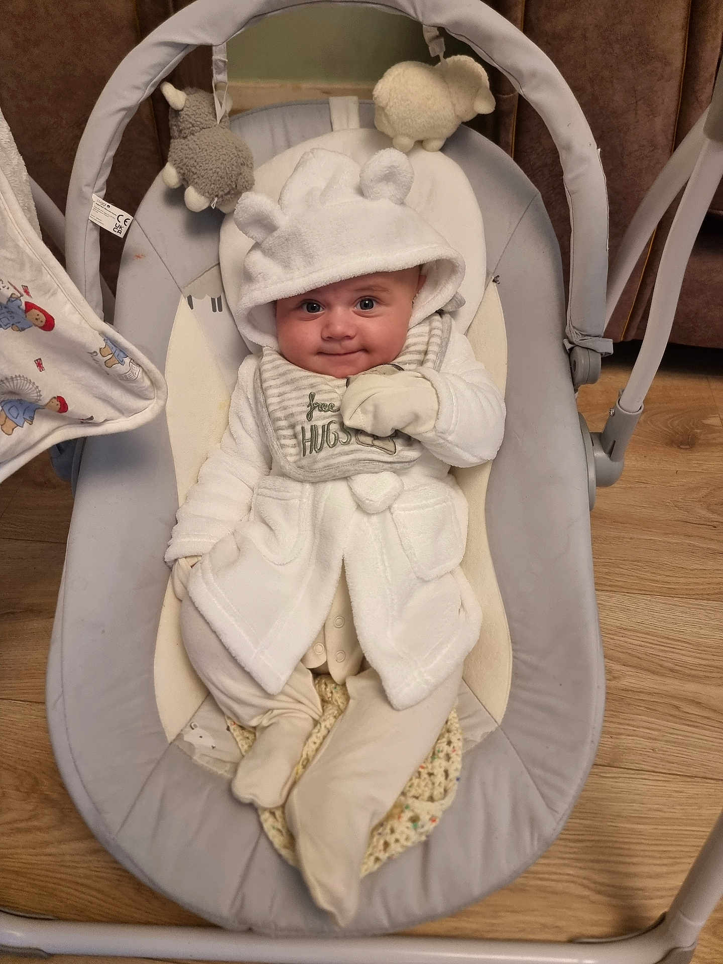 Ralph is registered to the contest to win money with this photo: baby, baby_bib, baby_rocker, child, clothing, cozy, cute, face, flooring, hood, indoor, infant, mittens, plush_toys, portrait, seated, smile, soft_toys, white_clothing, wood_floor
