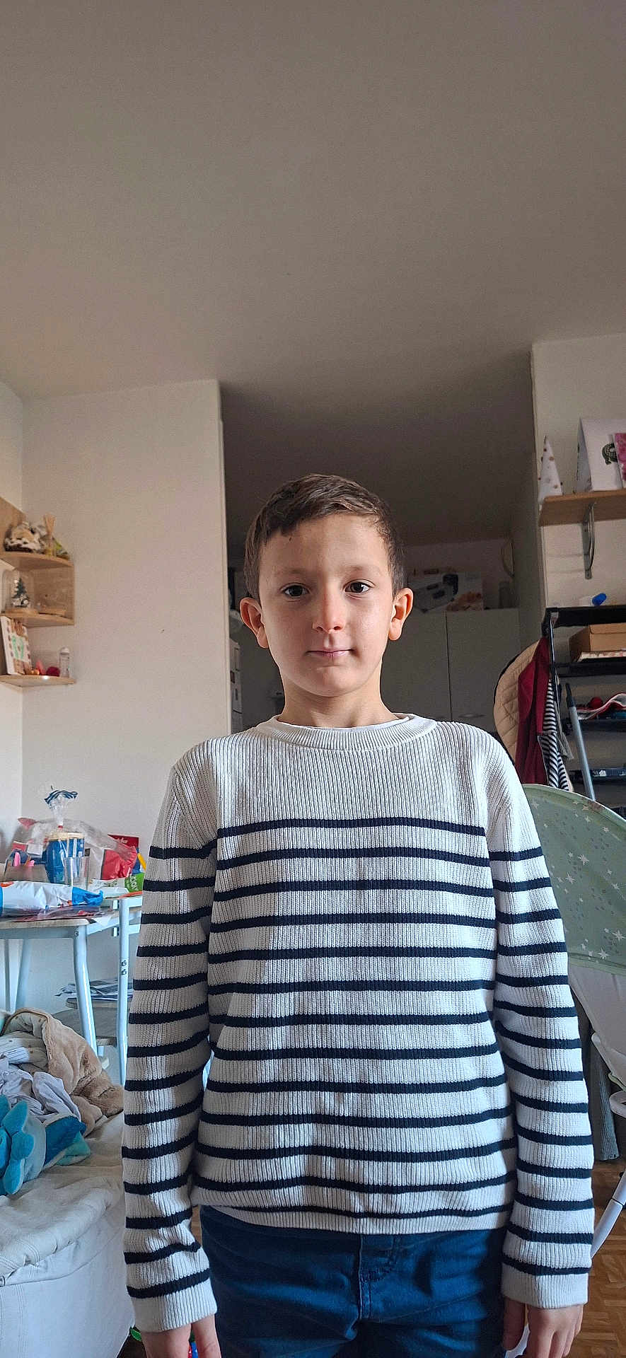 Thomas a rejoint le concours — aidez-le/la à gagner de superbes lots ! child, boy, striped_sweater, indoor, living_room, furniture, shelf, clothing, person, casual, portrait, standing, home, cabinet, toy, blanket, chair, floor, neutral_expression, short_hair