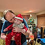 adult, baby, carpet, chair, christmas_tree, curtains, decorations, family, festive, holding, holiday, indoor, kiss, living_room, person, red_clothing, santa_hat, smile, table, warm_light
