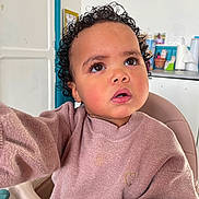 Hérondina participe au concours pour gagner de l'argent avec cette photo : toddler, child, curly_hair, pink_sweater, high_chair, indoor, kitchen, face, expression, looking_up, person, young_child, cozy, casual_clothing, home, portrait, cute, curiosity, seat, furniture