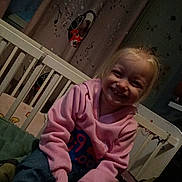 Laura a rejoint le concours — aidez-le/la à gagner de superbes lots ! child, smiling, blonde_hair, pink_hoodie, jeans, crib, bed, curtain, musical_notes, dog_graphic, indoor, happy, casual_clothing, young_child, person, portrait, sitting, cozy, room, furniture