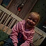 child, smiling, blonde_hair, pink_hoodie, jeans, crib, bed, curtain, musical_notes, dog_graphic, indoor, happy, casual_clothing, young_child, person, portrait, sitting, cozy, room, furniture