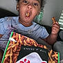 child, snack, pretzels, food, bag, curly_hair, mouth_open, hand, indoor, person, casual_clothing, couch, relaxed, funny_expression, brown_skin, earrings, window_blinds, bottle, sitting, home
