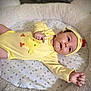 Albie is registered to the contest to win money with this photo: baby, child, yellow_onesie, hat, cushion, star_pattern, infant, cute, smiling, lying_down, cozy, soft_texture, indoor, portrait, happy, hand, face, skin, apparel, headwear