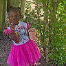 child, girl, pink_dress, unicorn, peace_sign, cup, outdoor, greenery, plants, wooden_posts, shoes, playful, smiling, nature, sunlight, young_child, casual, happy, standing, portrait