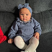 Stella is registered to the contest to win money with this photo: baby, child, infant, blue_clothing, headband, bow, tights, white_shoes, couch, gray_fabric, red_cushion, sitting, indoors, portrait, cute, face, hand, footwear, comfortable, young_child