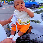 Ina participe au concours pour gagner de l'argent avec cette photo : baby, child, orange_shorts, white_shirt, giraffe_print, car, black_car, parking_lot, trees, cloudy_sky, adult_hands, outdoor, smiling, sitting, white_socks, person, vehicle, nature, daytime, cute