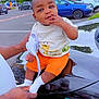 baby, child, orange_shorts, white_shirt, giraffe_print, car, black_car, parking_lot, trees, cloudy_sky, adult_hands, outdoor, smiling, sitting, white_socks, person, vehicle, nature, daytime, cute