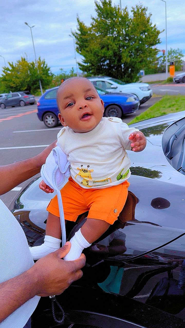 Ina a rejoint le concours — aidez-le/la à gagner de superbes lots ! baby, child, person, orange_shorts, white_shirt, giraffe_print, car, black_car, parking_lot, trees, greenery, outdoor, daylight, hands, holding, smiling, socks, road, vehicles, hood