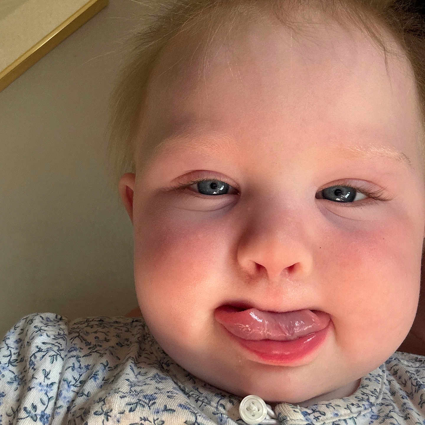 Rhea is registered to the contest to win money with this photo: baby, child, face, tongue, expression, close_up, cheeks, clothing, patterned_shirt, blue_eyes, skin, hair, button, indoors, portrait, person, cute, funny, infant, young_child