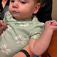 Brailee joined the competition — help win amazing prizes! toddler, child, baby, face, arm, hand, food, mouth, clothing, onesie, floral_pattern, person, holding, watch, skin, indoor, table, wood, adult, closeup