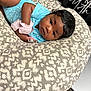 baby, infant, blue_onesie, cushion, patterned_fabric, pacifier, baby_bottle, indoor, carpet, curly_hair, face, looking, lying_down, soft_toys, baby_mittens, child, person, resting, cute, newborn