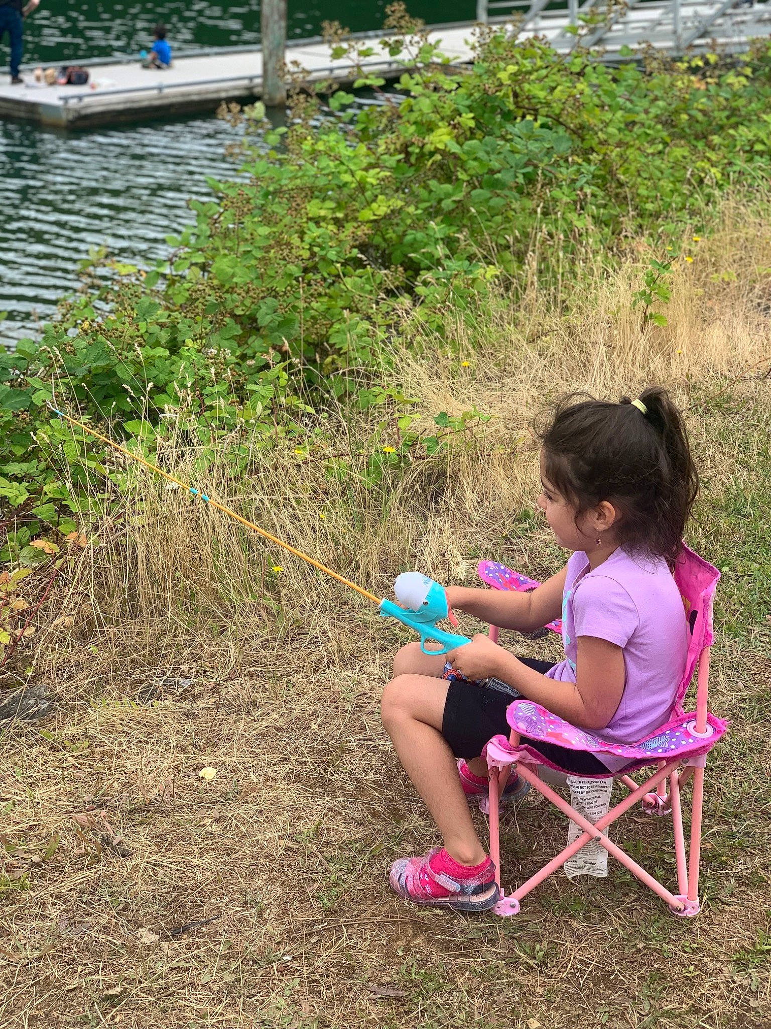 Mia is registered to the contest to win money with this photo: chair, child, fun, grass, grass_family, lake, leg, leisure, outdoor_furniture, people_in_nature, person, plant, recreation, sandal, shrub, sitting, soil, toddler, toy, tree