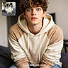 bed, beige, blue_eyed_cat, blue_eyes, blurred_background, casual_clothing, cat, cream, facial_expression, fluffy_cat, hair, hoodie, human, indoor, inset_photo, pet, portrait, surprised_expression, website_text, young_man