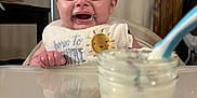 Nash joined the competition — help win amazing prizes! baby, bib, food, high_chair, smile, face, infant, table, spoon, jar, feeding, happy, cute, child, indoor, person, meal, messy, clothing, kitchen