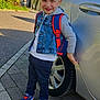 child, boy, denim_vest, backpack, sneakers, car, wheel, sidewalk, street, trees, daylight, outdoor, smile, casual_clothing, pavement, urban, building, blue_sky, hoodie, playful