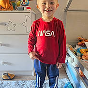 Maël a rejoint le concours — aidez-le/la à gagner de superbes lots ! child, boy, smiling, indoors, bedroom, toys, stuffed_animals, rug, barefoot, casual_clothing, red_shirt, blue_pants, furniture, drawer, bed, carpet, happy, person, playroom, portrait