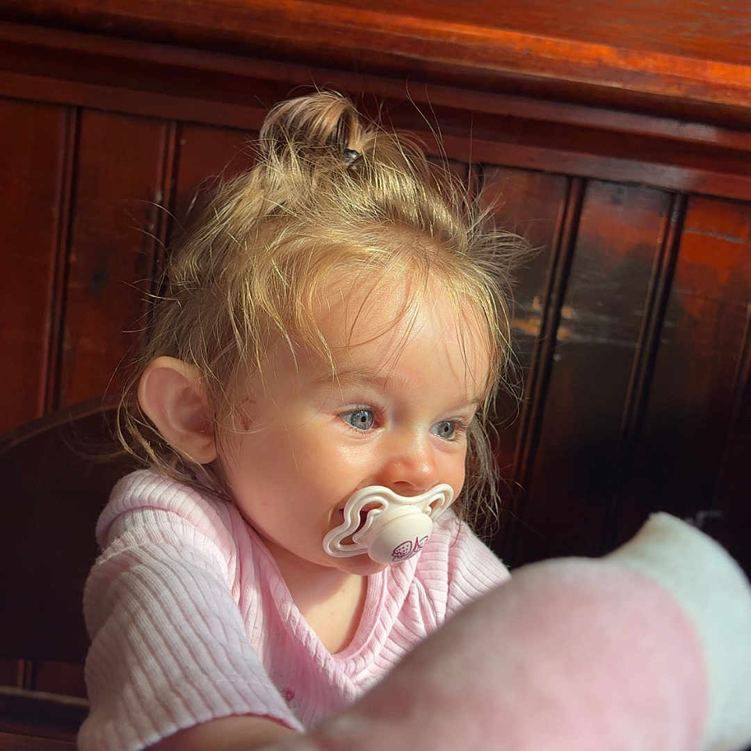 Isla-Robyn is registered to the contest to win money with this photo: baby, blond_hair, blue_eyes, child, curious, cute, fuzzy_toy, indoor, light, long_sleeve, natural_light, pacifier, person, pink_clothing, portrait, sitting, toddler, toy, wooden_background, young_child