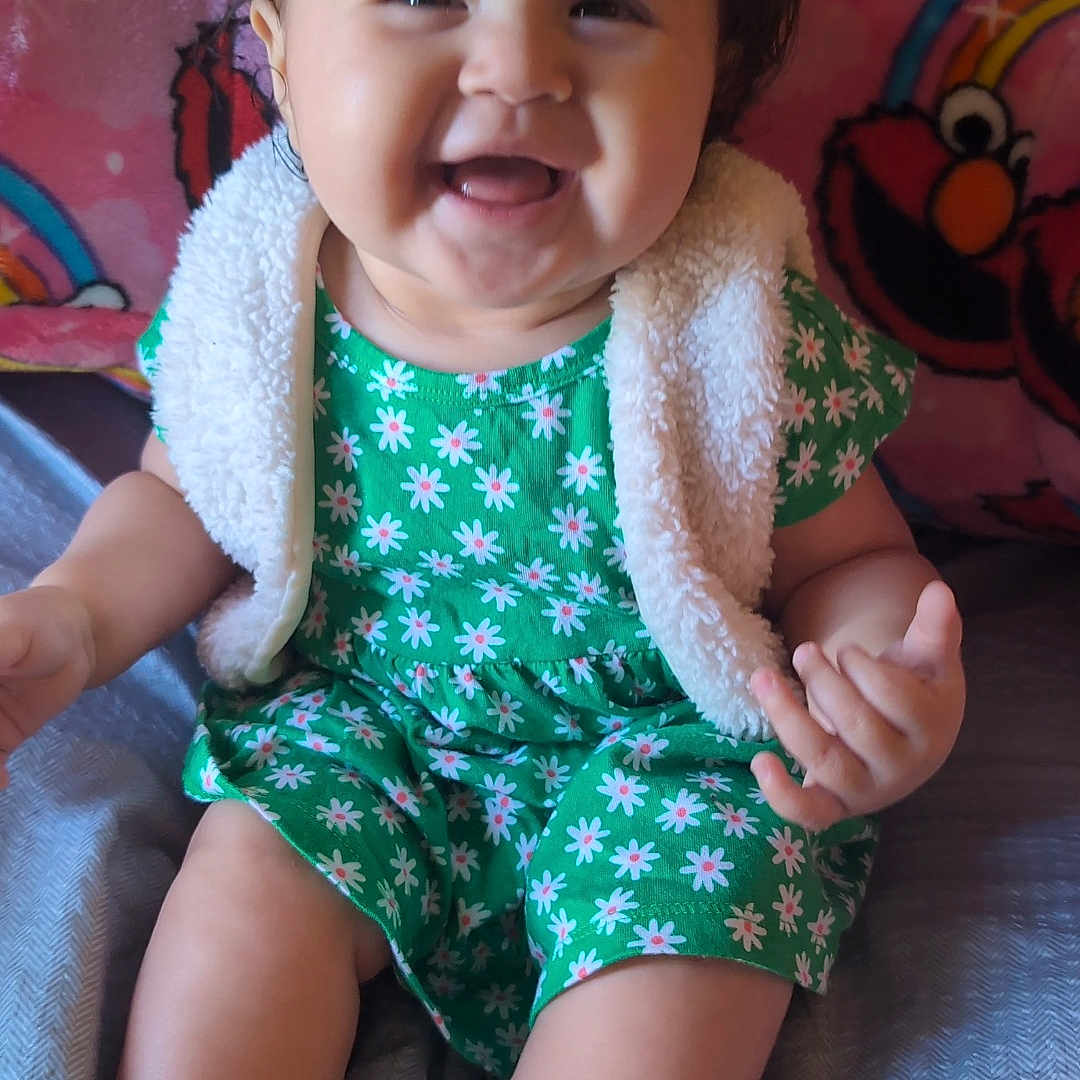 Fantasia is registered to the contest to win money with this photo: baby, bed, blanket, child, colorful, cute, elmo, face, floral_pattern, foot, green_dress, hair_bow, hand, happy, indoors, infant, portrait, sitting, smiling, vest