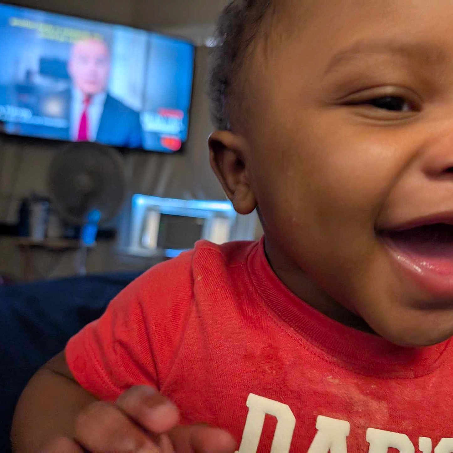 Azai is registered to the contest to win money with this photo: blurred_background, candid, casual, child, closeup, expression, face, furniture, hand, happy, home, indoor, living_room, person, portrait, red_shirt, skin, smiling, television, toddler