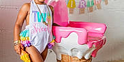 Stori Isabella is registered to the contest to win money with this photo: toddler, child, birthday, party, colorful_clothing, hair_accessories, ice_cream_cone, chair, wooden_floor, wall, indoor, portrait, smiling, shoes, socks, decoration, banner, playful, cute, fashion