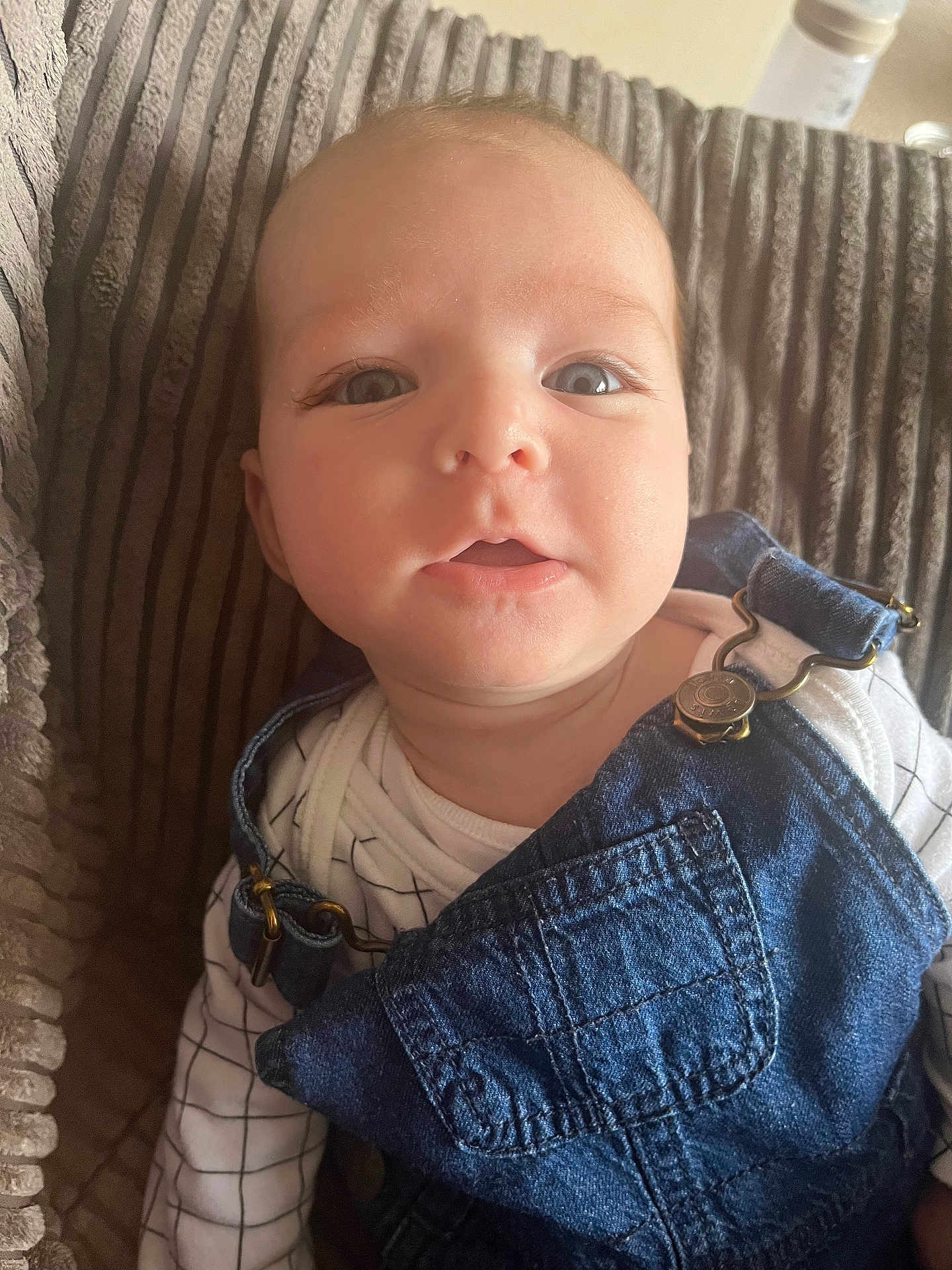 Theo is registered to the contest to win money with this photo: baby, child, denim, overalls, white_shirt, grid_pattern, cushion, texture, close_up, face, eyes, curious, soft_lighting, indoor, portrait, infant, clothing, person, skin, expression