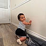 toddler, child, smiling, indoor, floor, wooden_floor, white_wall, tank_top, shorts, socks, baby, curious, playful, crawling, person, home, door, window_blinds, wall, flooring