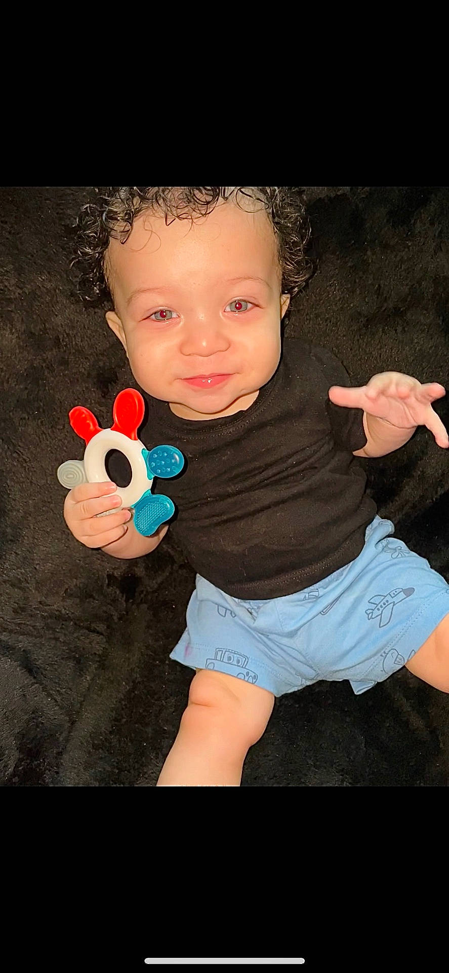 baby, black_shirt, blue_shorts, bright_eyes, casual_clothing, child, curly_hair, cute, face, fuzzy_surface, hand, indoor, infant, lying_down, person, playful, skin, smile, teething_toy, young_child