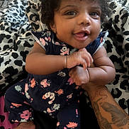 Sylea is registered to the contest to win money with this photo: baby, child, curly_hair, smiling, tattoo, hand, arm, bed, blanket, pattern, floral_clothing, infant, happy, indoors, person, cute, skin, foot, playful, holding