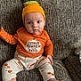 baby, child, infant, orange_hat, striped_shirt, pumpkin_outfit, cute, sitting, couch, home_interior, face, foot, hand, expression, text_on_clothing, patterned_pants, cozy, indoors, young_child, portrait