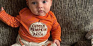 Jenson is registered to the contest to win money with this photo: baby, child, infant, orange_hat, striped_shirt, pumpkin_outfit, cute, sitting, couch, home_interior, face, foot, hand, expression, text_on_clothing, patterned_pants, cozy, indoors, young_child, portrait
